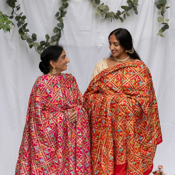 Two women in traditional phulkari standing against a white backdrop with decorative elements.

